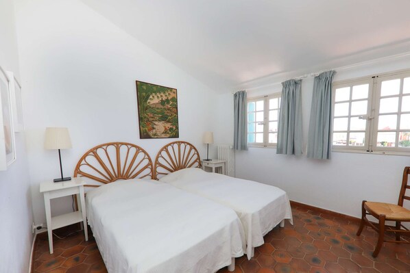 A room with two beds, a small table, a chair, a window with curtains, and a painting on the wall.