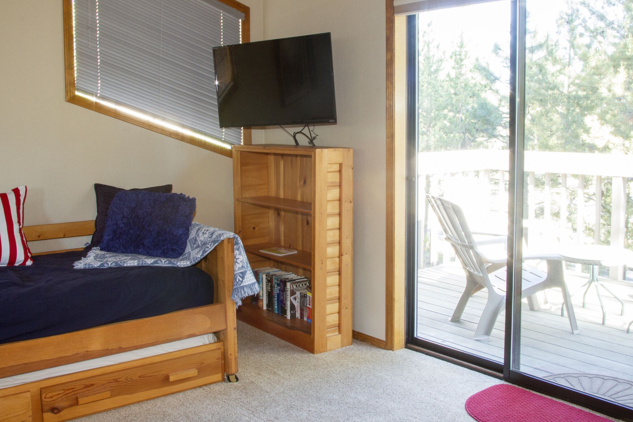 Une chambre avec un lit en bois, un téléviseur fixé sur une étagère en bois, une porte-fenêtre coulissante donnant sur un balcon et une petite bibliothèque.