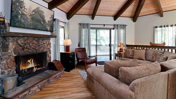 A cozy living room with a stone fireplace, wooden beams, a large sofa, and a view through a sliding glass door. - Maple Bear: Walk to Snow Summit! Hot Tub! Game Room! Pool Table! Air Hockey! Internet! Family Friendly! (Big Bear Lake)