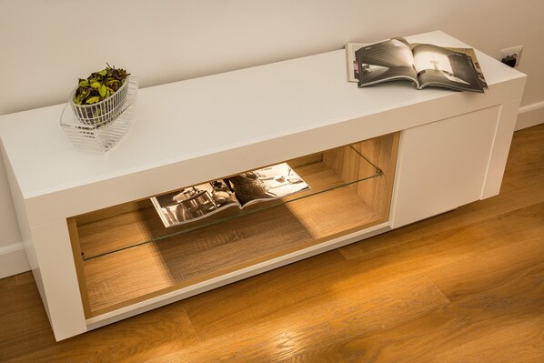 Une console blanche avec un tiroir à façade en verre, une petite plante et un magazine ouvert posés dessus.