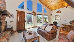 Un vaste salon doté d’un plafond en bois, d’une cheminée en pierre, d’un canapé en cuir et d’un coin repas avec une table et des chaises en bois.