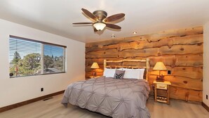Une chambre à coucher avec un lit, un mur en bois brut, un ventilateur de plafond, deux tables de chevet avec des lampes et une fenêtre avec des stores.