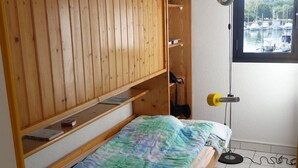 Una habitación pequeña y funcional con una cama, un armario de madera, un escritorio y una ventana con vista a barcos.