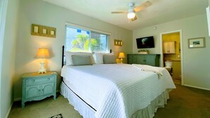 A bedroom with a bed, bedside lamps, a ceiling fan, a window with blinds, a television, and a bathroom visible through an open door. - 2nd House from the Beach with No Roads to Cross (Anna Maria)