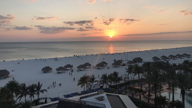 Une plage bordée de constructions aux toits de chaume et de palmiers, un ciel dégagé et le soleil se couchant sur l’océan.