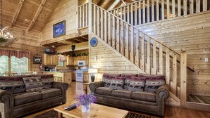A cozy cabin interior with a wooden staircase, two sofas with patterned cushions, a wooden dining table, and a kitchen area. - ERN849- Sweet Escape - Great Location! Close To Action! (Pigeon Forge)