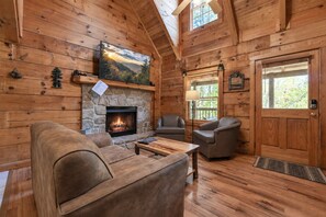 A cozy cabin interior with a stone fireplace, wooden walls, a comfortable sofa, and two armchairs.