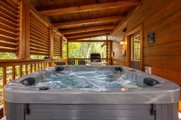 A wooden cabin with a hot tub in the center, surrounded by a deck and a forest view.