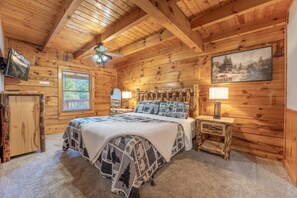 A cozy log cabin bedroom with a bed, two bedside lamps, a ceiling fan, a window with blinds, and a painting on the wall.