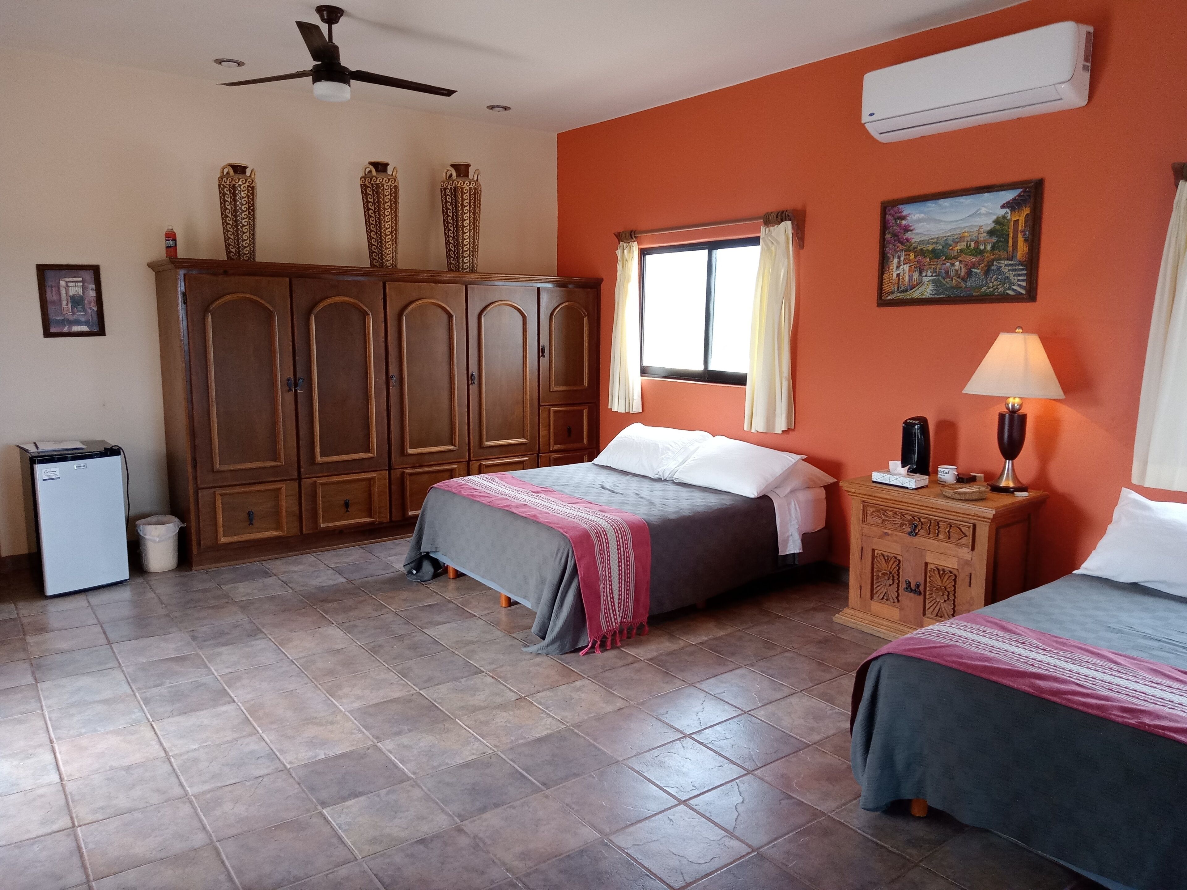 A hotel room with two beds, a wardrobe, a desk with a lamp, and a painting on the wall.