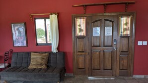 A room with a wooden door, a sofa with a cushion, a framed picture on the wall, and a window with curtains. - Hacienda de Palmas: Room #1 Grande with two queen size beds and sofa bed (La Ribera)