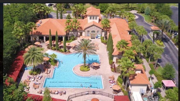Luchtbeeld van een resort met een groot zwembad, palmbomen en een gebouw met een rode pannendak.