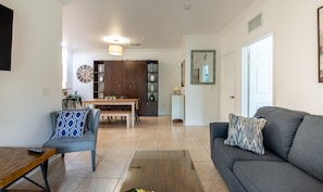 A living room with a grey sofa, a wooden coffee table, and a blue patterned armchair. A dining area with a wooden table and chairs is visible in the background.