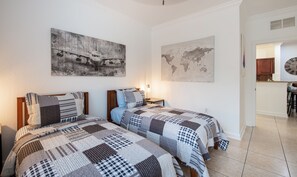 A bedroom with two beds, a nightstand, a wall map, and a kitchen area visible through an open door.