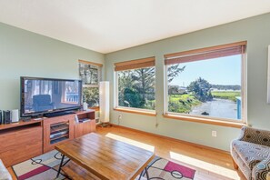 A living room with a TV, wooden furniture, large windows, and a view of a landscape.
