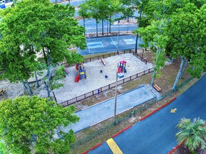 Children can play on the playground's slide, climbing frame, and circular play area surrounded by trees.