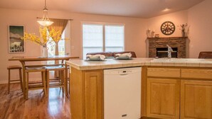 A kitchen with wooden cabinets, a dishwasher, a dining area with a table and chairs, a window with curtains, and a fireplace with a clock on the wall.