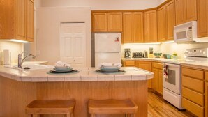 A kitchen with wooden cabinets, a white refrigerator, a microwave, and a stove. There are two wooden stools at the island.