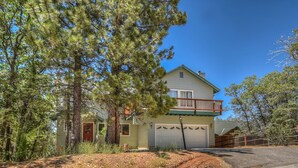 A two-story house with a garage, surrounded by trees and a driveway. - Mountain Crest Getaway: Mountain & Slope Views! Pool Table! Gas Fireplace! Cable! Pet Friendly! WiFi! (Big Bear City)