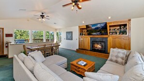 A spacious living room with a fireplace, a large TV, and a ceiling fan. - Mountain Crest Getaway: Mountain & Slope Views! Pool Table! Gas Fireplace! Cable! Pet Friendly! WiFi! (Big Bear City)