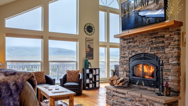 A spacious living room with a stone fireplace, large windows, leather furniture, and a view of mountains.