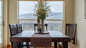 A dining area with a large wooden table, chairs, a chandelier, and a view through large windows.