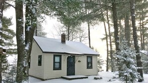 A small, single-story cabin with a snow-covered roof, surrounded by tall trees in a forest. - Lakefront Cabin #4 The Crappie (Presque Isle)