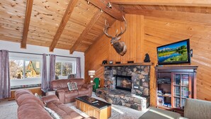 Ein gemütliches Blockhaus-Interieur mit Kamin aus Stein, Ledersofas, einem aufgehängten Hirschgeweih und einem Fernseher.
