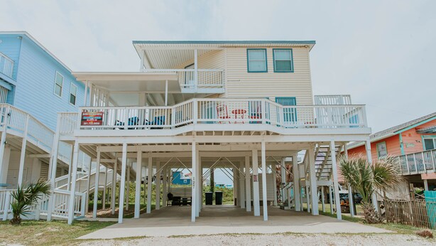 Een huis aan het strand met een overdekte veranda, meerdere balkons en een trap naar de bovenverdiepingen.