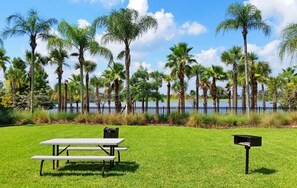 A picnic table with a bench, a trash bin, and a mailbox in a grassy area with palm trees and a body of water in the background. - Beautiful, Newly Renovated Condo! *Steps to Clubhouse/Pool* (Orlando)