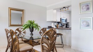 Un comedor con una mesa de vidrio rodeada de sillas de mimbre, un helecho en maceta y una cocina al fondo.