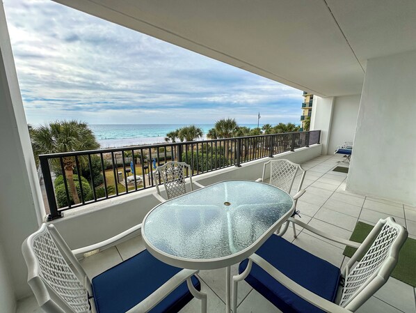 A balcony with a glass table and blue cushioned chairs, overlooking a beach and palm trees.