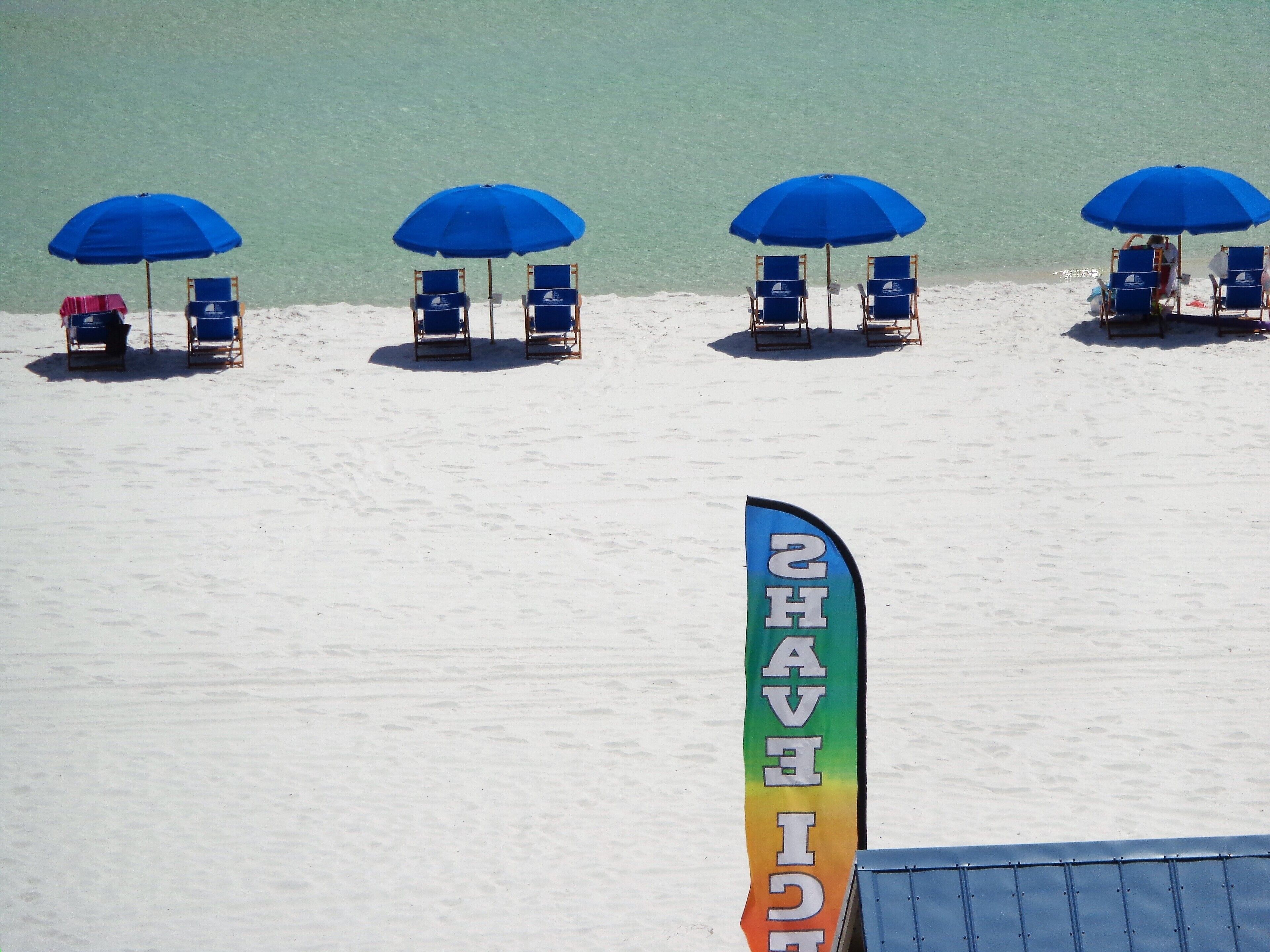 A beach with four blue umbrellas and lounge chairs. A flag with the text "AVE H" is visible.