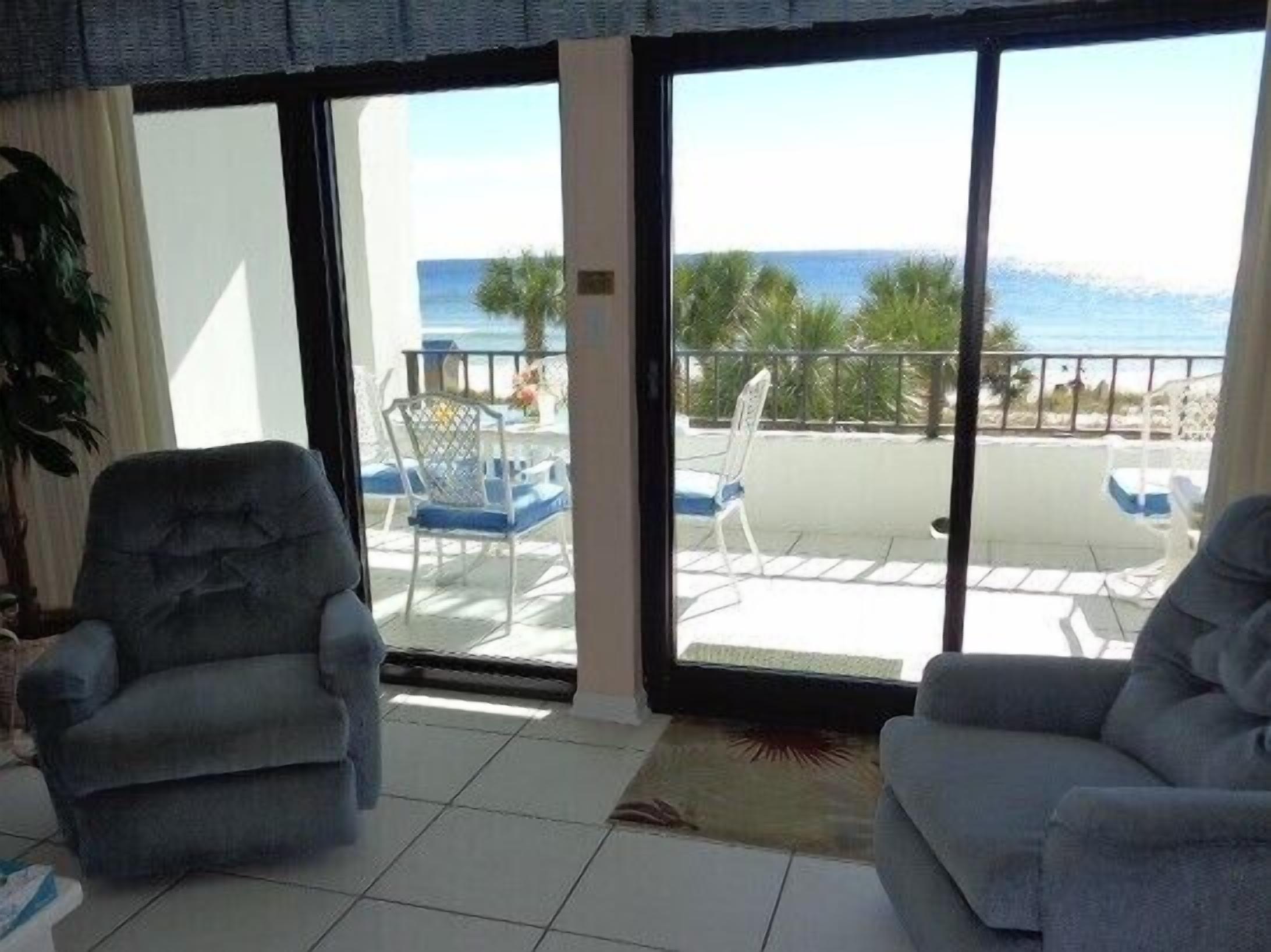 Une pièce avec deux fauteuils gris, une grande porte-fenêtre coulissante donnant sur un balcon avec vue sur la mer et les palmiers.