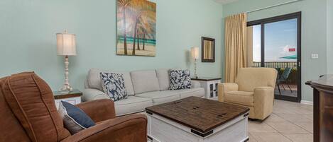 Un salon avec un mur bleu clair, un tableau représentant des palmiers, un canapé beige, un fauteuil marron, une table basse en bois et une porte en verre donnant sur un espace extérieur.