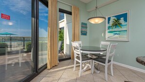 A room with a glass door leading to a balcony, a round glass table with white chairs, a hanging light fixture, and a framed picture of a beach scene. - *Ground Floor* 1BR @ Phoenix VII (Orange Beach)