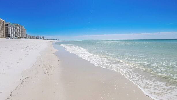 A beach with a clear sky, a sandy shore, and a row of high-rise buildings in the background.