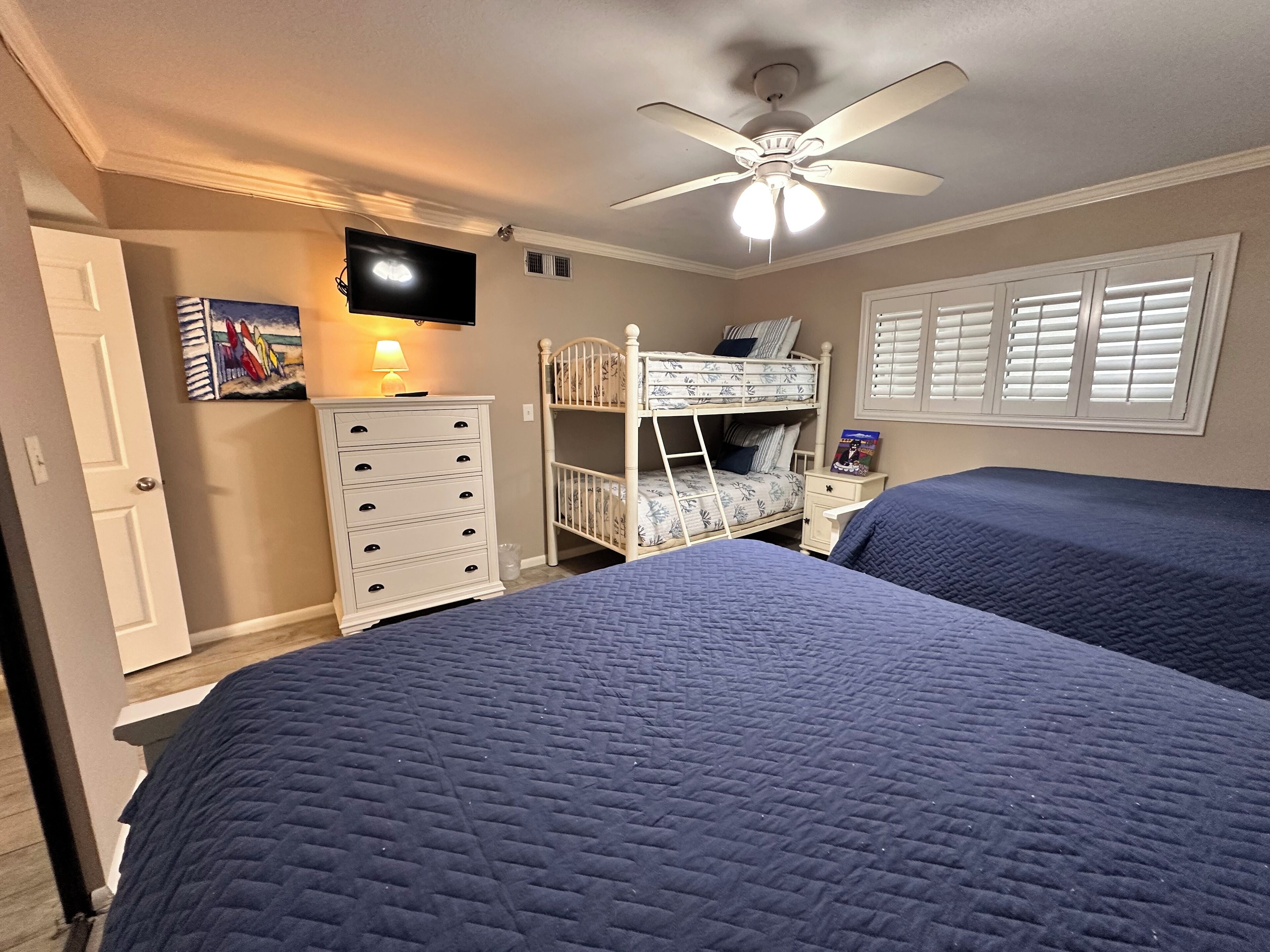 A bedroom with two beds, a ceiling fan, a TV, a dresser, and a window with shutters.