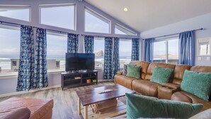 A living room with a leather sofa, a coffee table, and a TV on a stand. There are large windows with curtains, and a view of the beach is visible. - Seaside Splendor - large home, fantastic views very, close to beach (Pacific City)