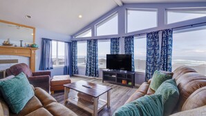 A spacious living room with a large window, a flat-screen TV, leather sofas, and a wooden coffee table. - Seaside Splendor - large home, fantastic views very, close to beach (Pacific City)