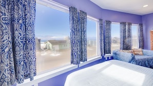 A bedroom with a large bed, a window with blue curtains, and a view of houses outside. - Seaside Splendor - large home, fantastic views very, close to beach (Pacific City)