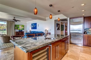A modern kitchen with a large island, pendant lighting, and a stainless steel refrigerator.