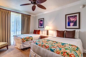 Quarto de hotel com duas camas, um ventilador de teto, uma mesa de cabeceira com abajur e dois quadros florais emoldurados na parede.