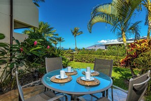 A round table with four white cups and saucers, surrounded by wicker chairs, set on a blue glass table in a garden with palm trees and a view of a mountain.