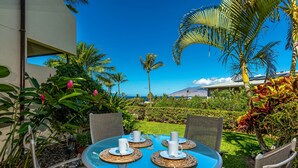 Una mesa redonda con cuatro tazas y platillos blancos, rodeada de sillas de mimbre, sobre una mesa de vidrio azul en un jardín con palmeras y vistas a una montaña.