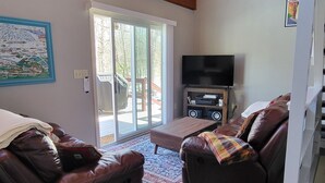 A spacious living room with leather sofas, a flat-screen TV, and a large window with a view of a deck. - Hike, ski, summer fun in our all season home minutes to Okemo. Dogs welcome! (Mt Holly)
