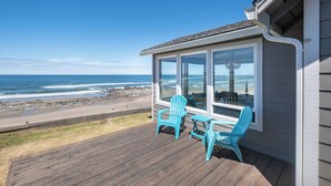 Eine Holzterrasse mit zwei blauen Adirondack-Stühlen und einem Tisch, mit Blick auf einen Strand mit felsigem Ufer und Meereswellen.