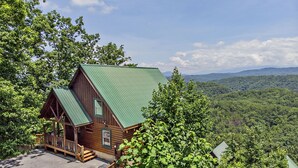 Une cabane en bois avec un toit vert, une véranda et une vue sur une chaîne de montagnes boisées.