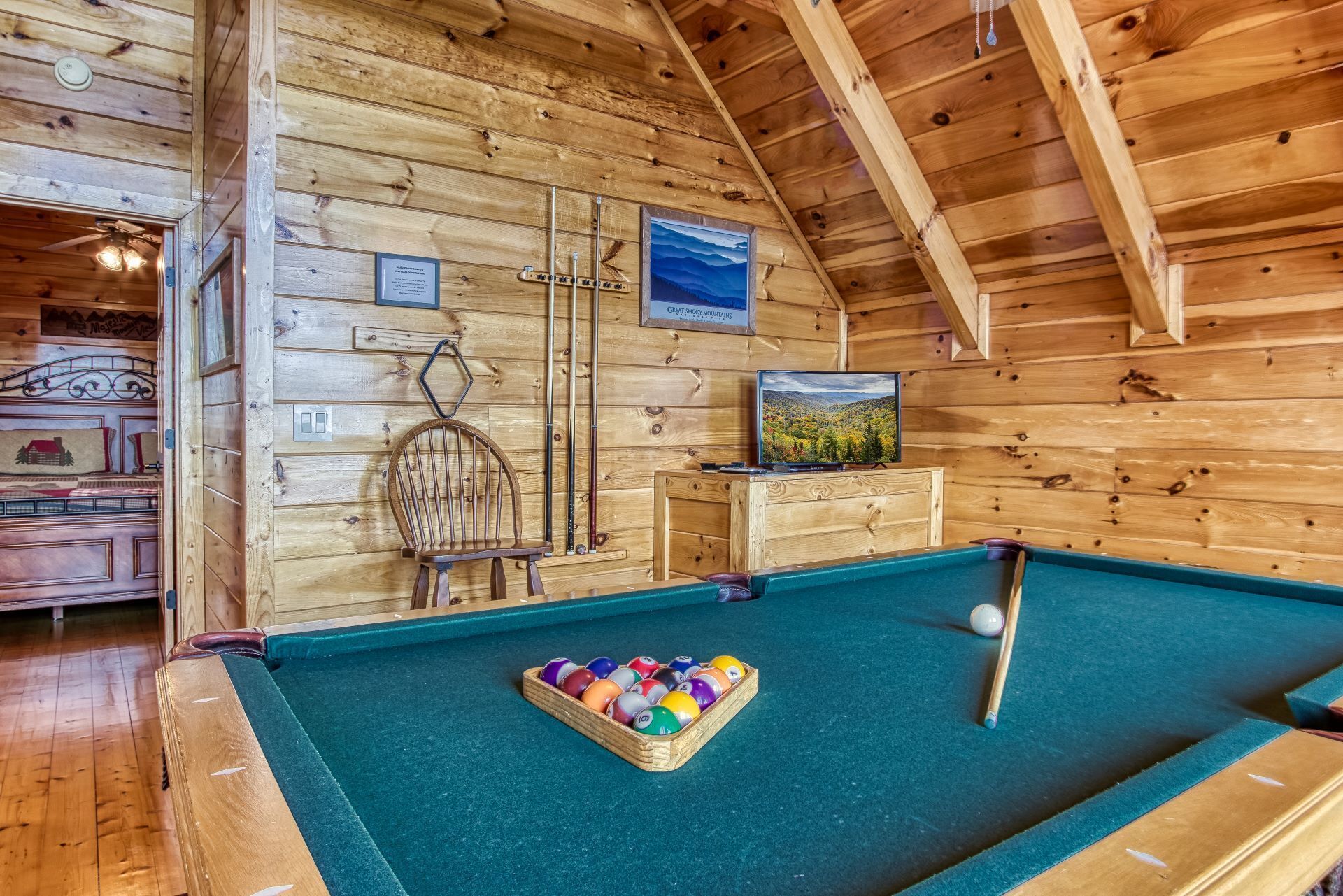 A pool table with a cue ball and a box of billiard balls, wooden walls, a mounted television, and a framed picture of mountains.
