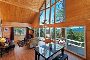 A spacious cabin with a high ceiling, large windows offering mountain views, a cozy seating area, and a dining table.